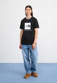 Camiseta de algodón negra con logo y texto blancos, combinada con unos vaqueros azul claros y zapatillas marrones. El modelo está de pie sobre una superficie azul clara.