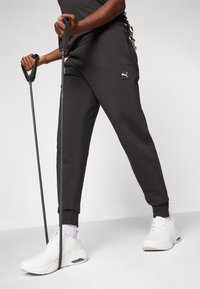 Schwarze Jogginghosen mit elastischen Bündchen, versehen mit einem kleinen Logo-Patch, kombiniert mit weißen Sportschuhen. Die Person hält Trainingsstöcke.