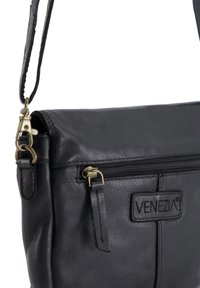 Sorte læder crossbody taske med en struktureret overflade, der har en frontlomme med lynlås, et mærke med logo, og en metalspænde på en lang rem.