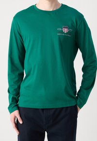 Długorękawowa zielona koszula wykonana z bawełny, z okrągłym dekoltem i małym haftowanym logo na piersi. Gładka tekstura, luźny krój.