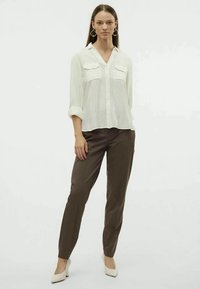 Camicia bianca strutturata con due tasche sul petto, abbinata a pantaloni marroni e tacchi bianchi. Modello in piedi davanti a uno sfondo neutro.