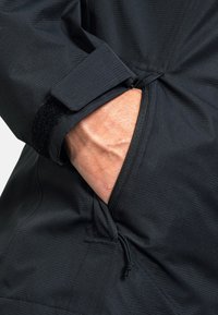 Schwarze Jacke mit strukturiertem Finish, ausgestattet mit einem Klettverschluss am Ärmel und einer seitlichen Tasche mit Reißverschluss.