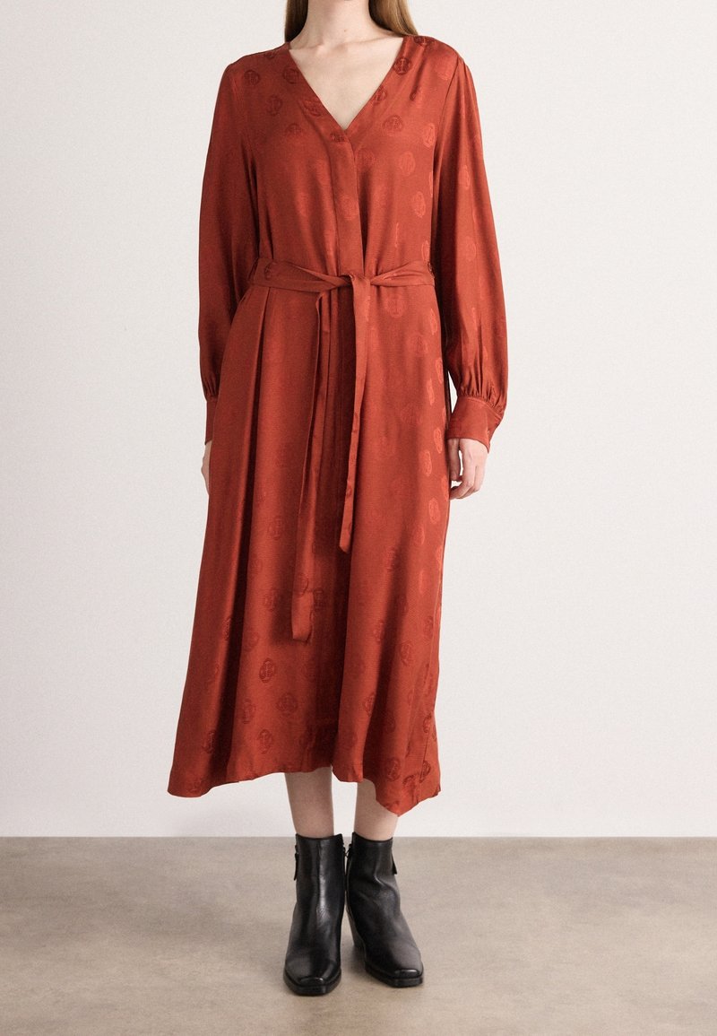 Femme portant une robe midi à manches longues couleur rouille avec ceinture nouée à la taille et motif subtil, associée à des bottines noires sur un sol gris.