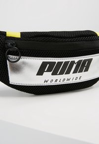 Sac banane en mesh noir avec un patch argenté indiquant le logo "PUMA WORLDWIDE" et une fermeture éclair noire avec un curseur rond sur fond blanc.