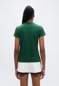 Άτομο με μακριά μαλλιά δεμένα πίσω, που φοράει πράσινο κοντομάνικο πουκάμισο και λευκό σορτς με σκούρο περίγραμμα, στέκεται γυρισμένο με την πλάτη σε απλό φόντο.