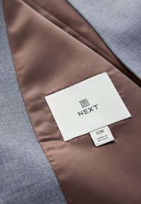 Tissu gris texturé avec une doublure en satin marron et une étiquette blanche portant la mention "NEXT" et la taille 40R. Mention également "FABRIQUÉ AU VIETNAM".