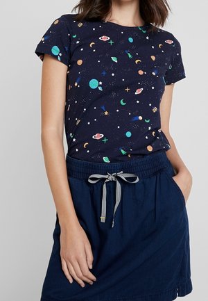 Kobieta nosząca granatowy t-shirt z kolorowymi nadrukami planet i gwiazd oraz ciemnoniebieską spódnicę ze sznurowadłem w paski.