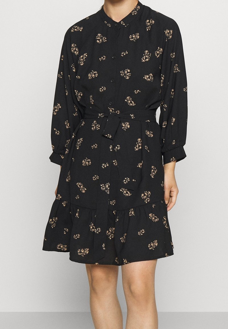 Femme portant une robe noire à manches longues avec de petits imprimés floraux beige, boutonnée devant, ceinture à la taille et ourlet à volants.