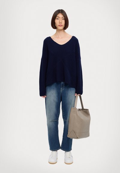 Maglione a costine blu navy con scollatura a V, taglio oversize; abbinato a jeans baggy azzurri chiari e sneakers bianche; tiene in mano una borsa tote beige.