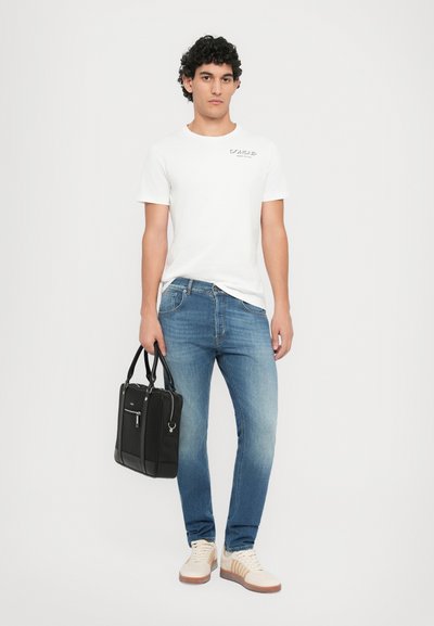 Λευκό βαμβακερό μπλουζάκι με λογότυπο, ανοιχτό γαλάζιο τζιν παντελόνι, μπεζ sneakers και μια μαύρη δομημένη τσάντα ώμου. Χωρίς σχέδια ή υφές ορατές.