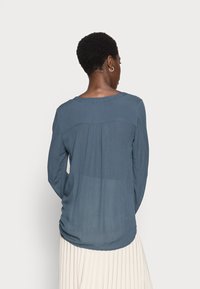 Blouse à manches longues en vert émeraude, avec un col rond, une texture douce et un détail plissé à l'arrière, associée à une jupe légère plissée.