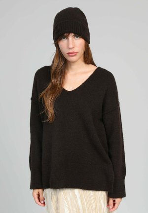 Jeune femme aux longs cheveux bruns portant un bonnet en tricot noir, un pull à col en V noir et une jupe texturée de couleur claire sur un fond uni.