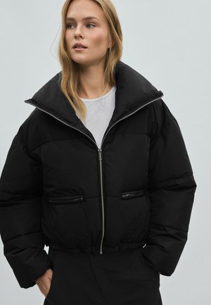 Frau mit blondem Haar, die eine schwarze Steppjacke mit hohem Kragen über einem weißen Hemd trägt, Hände in den Taschen, schaut leicht zur Seite.
