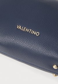 Temno modra usnjena denarnica z teksturirano površino, ki ima zlatotone logo "VALENTINO" in mesingaste dodatke. Vidne podrobnosti šivanja.