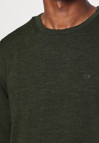 Mörkgrön stickad tröja med rund halsringning, strukturerat tyg och en liten "CK"-logga broderad i det nedre hörnet.