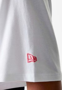Witte stof met een klein rood geborduurd logo nabij de zoom, waarbij een deel van de arm van een persoon die het kledingstuk draagt, te zien is.
