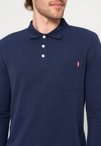 Άνδρας φοράει μπλε polo με μακριά μανίκια, τρία λευκά κουμπιά και τσέπη στο στήθος με μικρή κόκκινη, λευκή και μπλε λωρίδα.