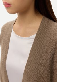 Brun kofta med texturerat stickat mönster, med v-ringning och öppen front, bärs över en vit topp. Mjuk och len tyg.