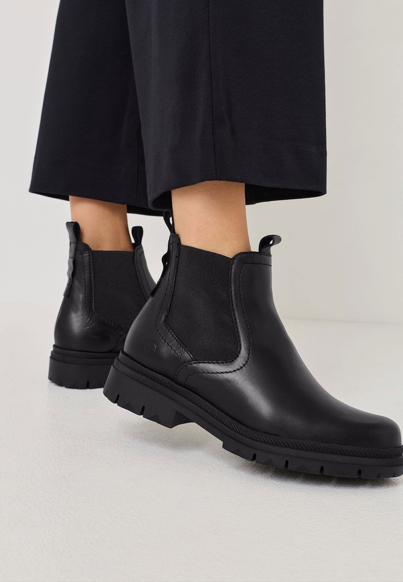 Botoșei din piele neagră până la gleznă, cu panouri elastice pe laterale, vârfuri rotunde și tălpi din cauciuc texturat. Accentele includ linii mici pentru tragere la spate.