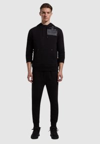 Felpa nera con cappuccio, tasca frontale e dettaglio del logo, abbinata a pantaloni jogger neri e sneakers nere.