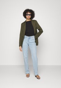Olivově zelená blazer, černý top, světle modré džínsy s vysokým pasem a černé sandály na podpatku. Modelka pózuje s kudrnatými vlasy a slunečními brýlemi.