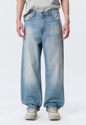 Persona che indossa jeans larghi, sbiaditi azzurri e sneakers beige, in piedi davanti a uno sfondo chiaro e uniforme.