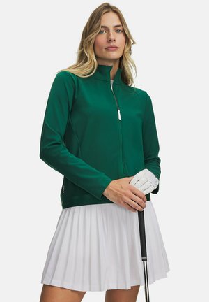 Vrouw met een groen jack met ritssluiting en een witte geplooide rok die een golfclub vasthoudt met een witte handschoen tegen een effen achtergrond.