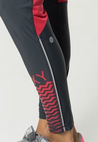 Czarne i czerwone legginsy sportowe z wzorem w zygzak, gładka faktura oraz odblaskowe akcenty po bokach. Widoczny detal logo.