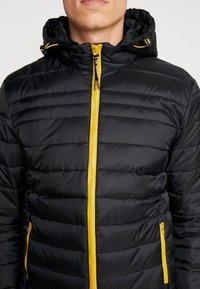 Homme portant une veste matelassée noire avec une fermeture éclair jaune et des cordons à la capuche, montré du cou à la taille sur un fond uni.