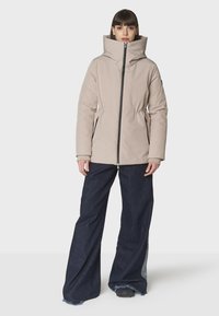 Mujer vestida con chaqueta de plumas beige de cuello alto con cremallera negra, jeans oscuros de pierna ancha y zapatos negros, de pie frente a un fondo blanco.
