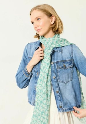 Nainen pukeutunut vaaleansiniseen farkkutakkiin valkoisen mekon päällä, säätää vaaleanvihreää huivia, jossa on valkoinen lehtikuvio, katsoo sivulle.