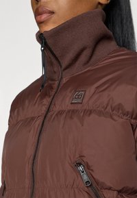Casaco puffer castanho com um colarinho alto com nervuras, fecho frontal com zíper e bolso lateral. Apresenta um patch com o logótipo no peito. Textura suave.