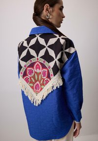 Giacca blu trapuntata con tessuto nero a motivo floreale sul retro, dotata di dettagli frangiati. Il colletto e le maniche sono di un blu uniforme.