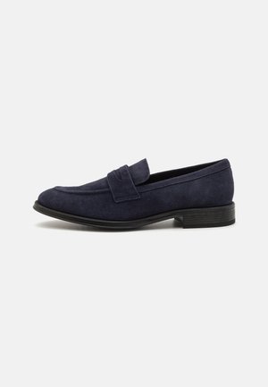 Mocassins en daim bleu marine avec un bout arrondi, dotés d'une bride décorative et d'un petit talon noir, texture lisse et design minimaliste.