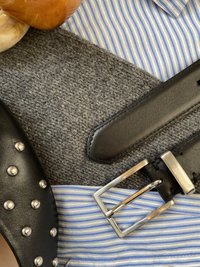 Juodas odinis diržas su sidabriniais vinutėmis, stačiakampiu metaliniu sagtimi, padėtas ant pilko tekstūruoto paviršiaus, šalia mėlynai dryžuotos medžiagos.