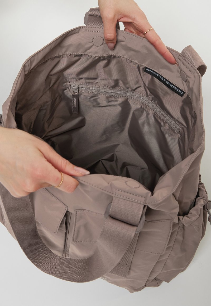 Sac fourre-tout en tissu recyclé de couleur taupe atténuée, avec une poche zippée et des compartiments intérieurs. Bretelle épaulière épaisse et réglable pour un soutien optimal.
