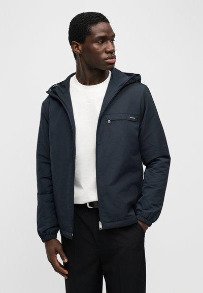 Άνδρας που φοράει σκούρο μπλε μπουφάν με κουκούλα πάνω από λευκό πουκάμισο και μαύρο παντελόνι, κοιτάζει δεξιά με το ένα χέρι στην τσέπη.