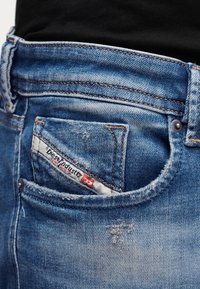 Κοντινό πλάνο μπλε τζιν τζιν με ετικέτα Diesel Industry στην μικρή μπροστινή τσέπη και ελαφρά φθαρμένες λεπτομέρειες.