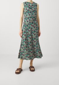 Vestido floral con cuello drapeado, base verde y patrones en rojo y rosa. Falda hasta la rodilla; combinado con sandalias de tira trenzadas en color marrón.