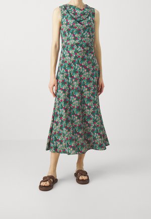 Robe fleurie avec un col drapé, base verte et motifs rouges et roses. Jupe jusqu'aux genoux ; associée à des sandales brunes à enfiler tressées.