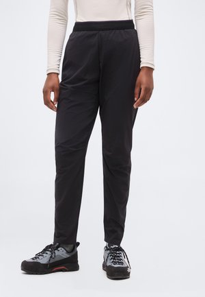 Asmuo dėvi juodus siaurėjančius kelnes ir pilkus juodus žygių batus, stovi priešais paprastą šviesią foną.