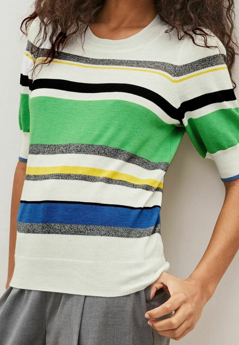 Personne portant un pull à manches courtes blanc avec des rayures horizontales vertes, bleues, noires, jaunes et grises, associé à un pantalon gris.