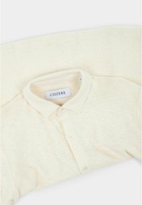 Crème-kleurig shirt van een gestructureerde stof, plat gevold. Beschikt over een kraag, knopenlijst en een label met de tekst "CISZERE."