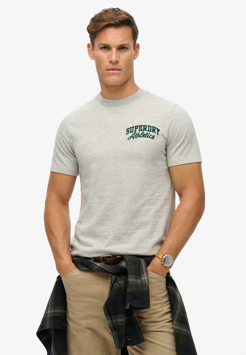 Graues T-Shirt mit kurzen Ärmeln und dunkelgrünem "Superdry Athletics" Logo auf der linken Brust. Das Modell trägt beige Hosen und ein kariertes Hemd, das in der Taille gebunden ist.