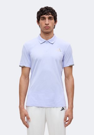 Mand står og vender fremad iført en lyse-lilla Adidas polo og hvide bukser med Adidas-logo, mod en ensfarvet baggrund.