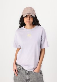 Tricou din bumbac de culoare violet deschis, cu mâneci scurte și decolteu rotund, având un grafic mic cu o palmier în galben, asortat cu o șapcă bej.