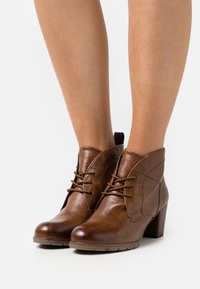 Braune Lederstiefeletten mit Schnürdesign, mit einer geschwungenen Schnittführung, strukturierten Oberflächen und einem gestapelten Absatz.