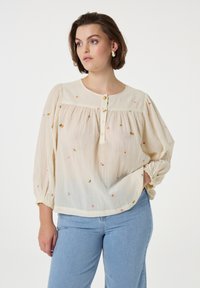 Cremfarbene Bluse mit floralem Stickmuster. Hat einen runden Halsausschnitt, drei Knöpfe und elastische Bündchen. Leichtes, durchscheinendes Material.