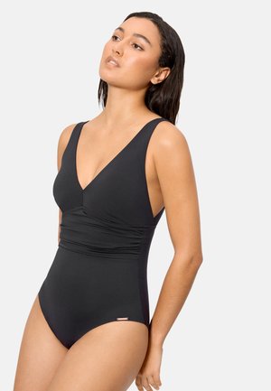 Mujer con cabello largo y oscuro, vistiendo un traje de baño negro de una pieza con escote en V y detalle fruncido en la cintura, posando frente a un fondo claro y liso.