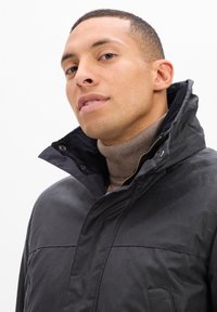 Mężczyzna w czarnej kurtce z podniesionym kołnierzem na brązowym golfie, pewnie spoglądający w kierunku kamery na jednolitym białym tle.